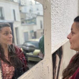 Yaëlle Dorison vor einem Spiegel und schaut sich selbst an. Sie trägt eine dunkelrote Jacke und im Hintergrund sind Häuser zu sehen