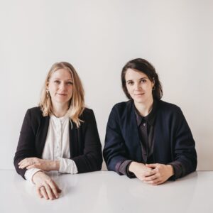 Portrait von OODD Studios Susanne Ostwald und Magdalena Sophie Orland sitzend an einem Tisch in dunklen Klamotten