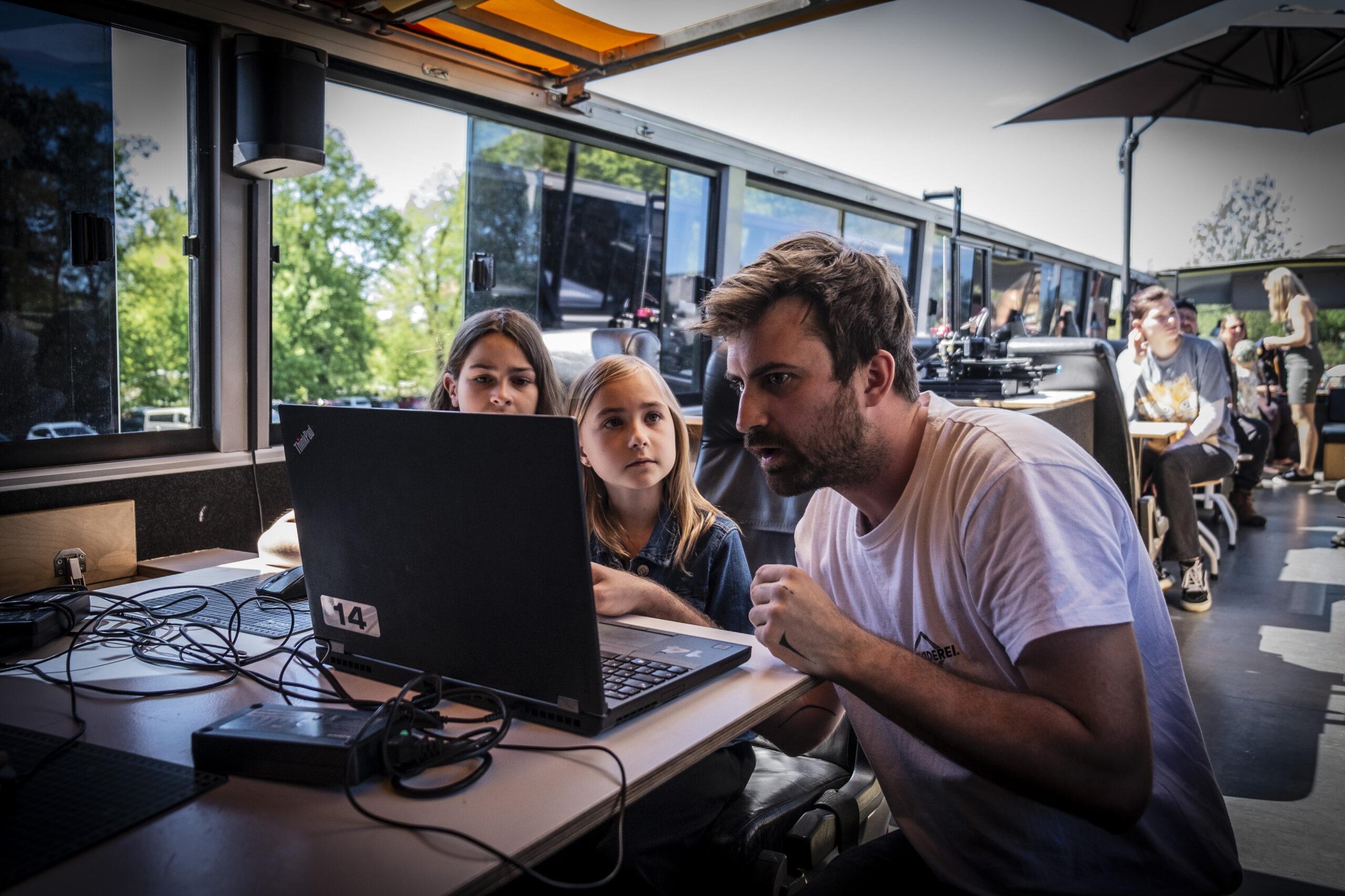 zwei Mädchen und ein Mann sitzen an einem Laptop im Bus des Fabmobil mit offenem Dach