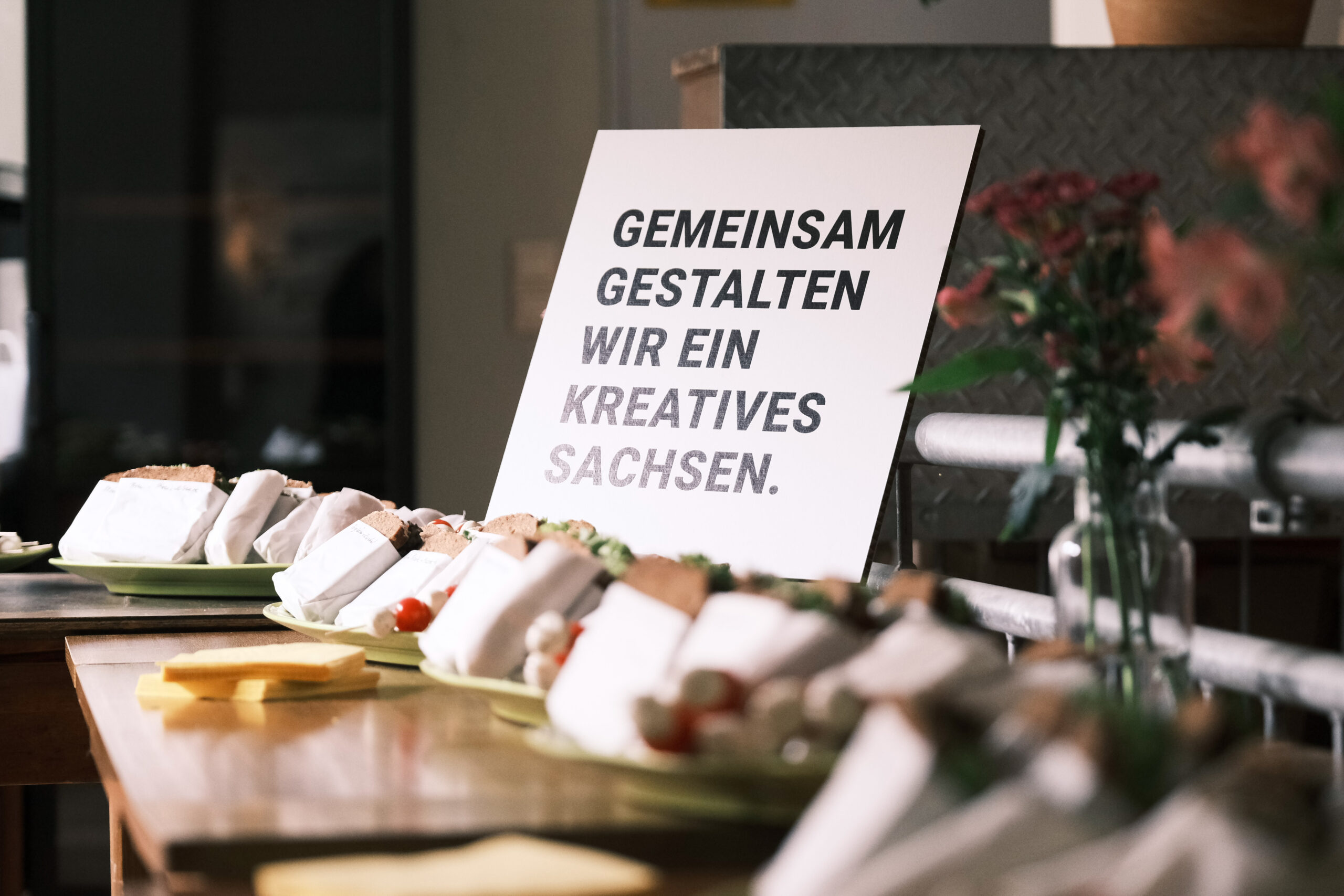 Der Schriftzug Gemeinsam Gestalten wir ein Kreatives Sachsen auf einem Karton, im Vordergrund belegte Brote als Buffet aufgebaut