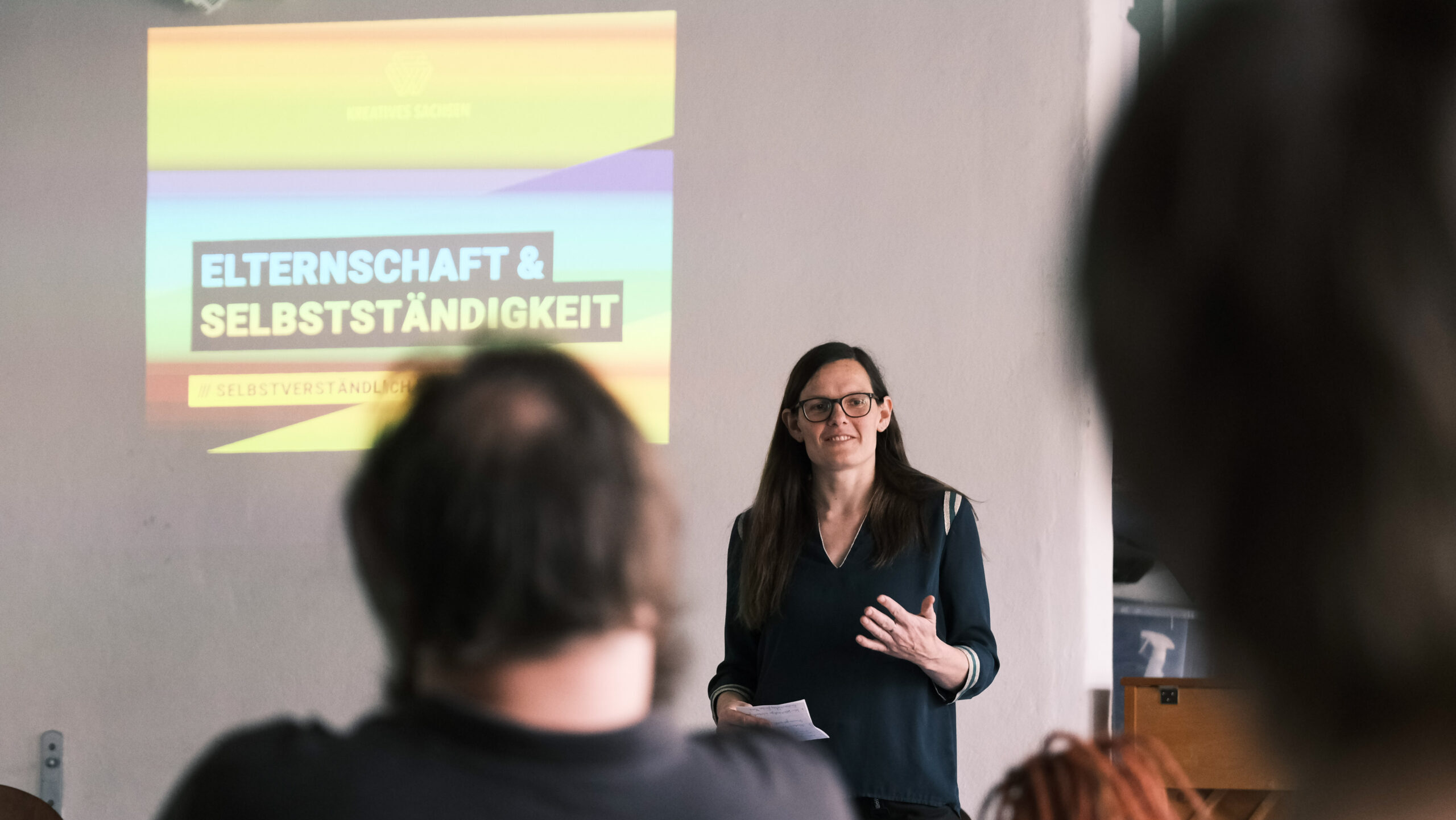 Frau spricht vor Präsentation im Vordergrund sitzen Personen von hinten sichtbar