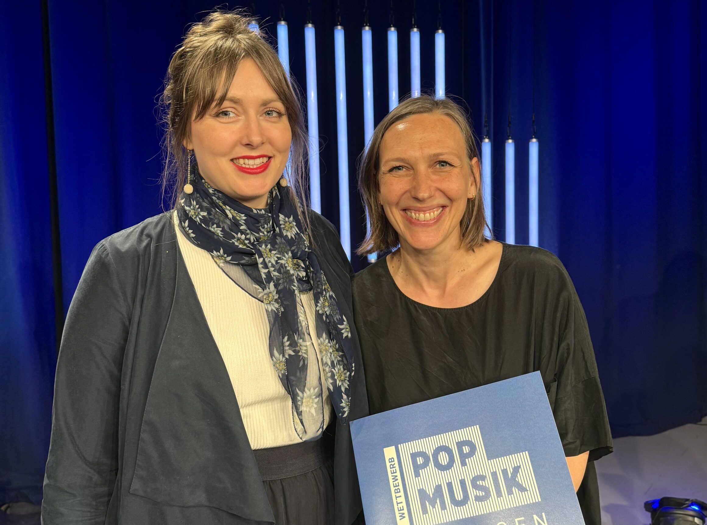 Zwei Frauen stehen auf einer blau beleuchteten Bühne und lachen in die Kamera. Eine Frau hält eine Urkunde mit der Aufschrift: Wettbewerb Pop Musik Sachsen 2024.