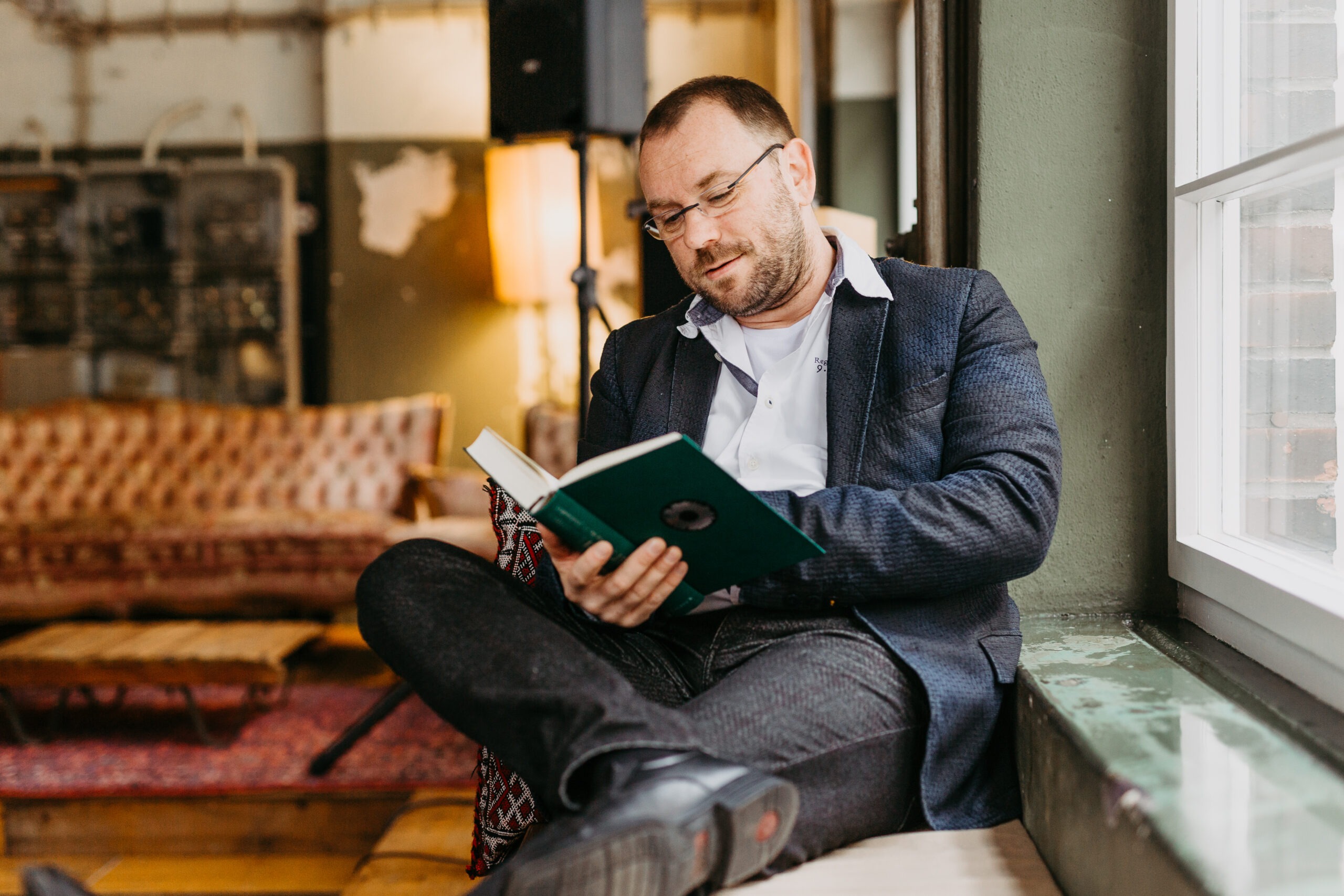 Mann mit kurzen Haaren und Brille liest in einem Buch. Er hat das rechte Bein übergeschlagen und sitzt an einem Fenster.