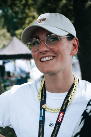 Frau mit Brille, kurzen Haaren und einer weißen Käppi. Sie schaut in die Kamera. Sie trägt ein weißes Shirt und ein schwarzes Schlüsselband und lächelt in die Kamera.