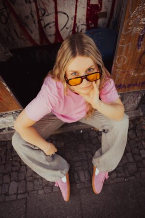 Frau in rosa Shirt mit blonden Haaren. Sie trägt eine orangene Sonnenbrille und schaut von unten in die Kamera.