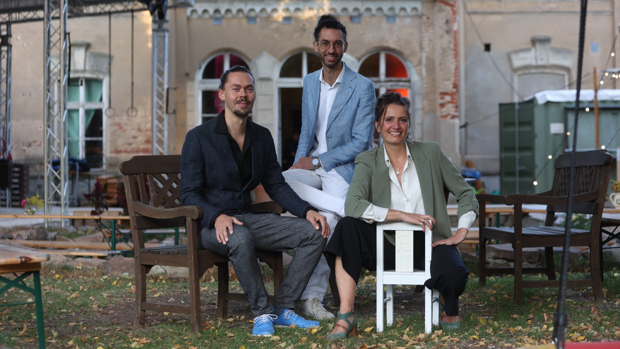 Zwei Männer und eine Frau sitzen auf Stühlen vor einem alten Gebäude, alle lächeln