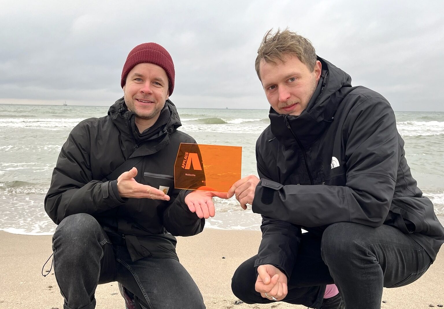 Zwei weiße Männer in der Hocke an einem Strand, sie halten gemeinsam einen orangenen Preis aus Glas in der Hand mit der Aufschrift APPLAUS.