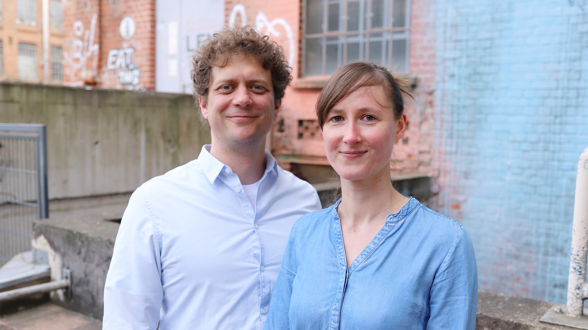 Zwei weiße Personen vor einer groben Mauer. Links ein Mann mit Locken und weißem Hemd, rechts eine Frau mit Seitenscheitel und Jeansbluse. Beide lächeln.