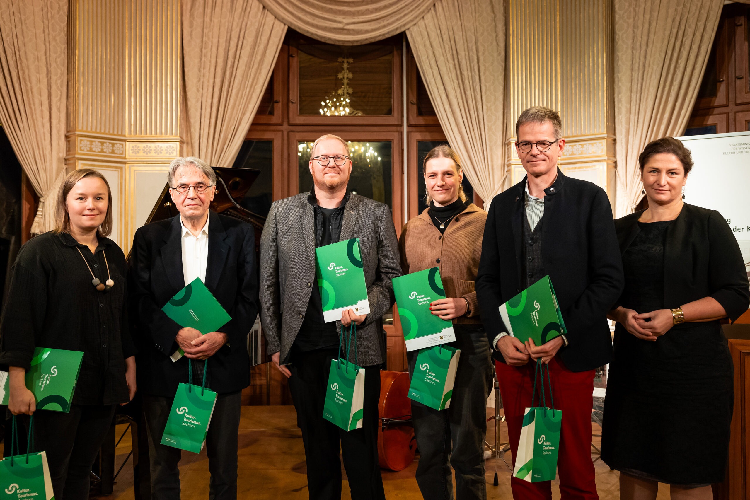 Sechs weiße Menschen, drei Frauen und drei Männer, alle schick gekleidet. Fünf Menschen halten eine grüne Urkunde und grüne Taschen in der Hand mit der Aufschrift: Kultur Tourismus Sachsen.