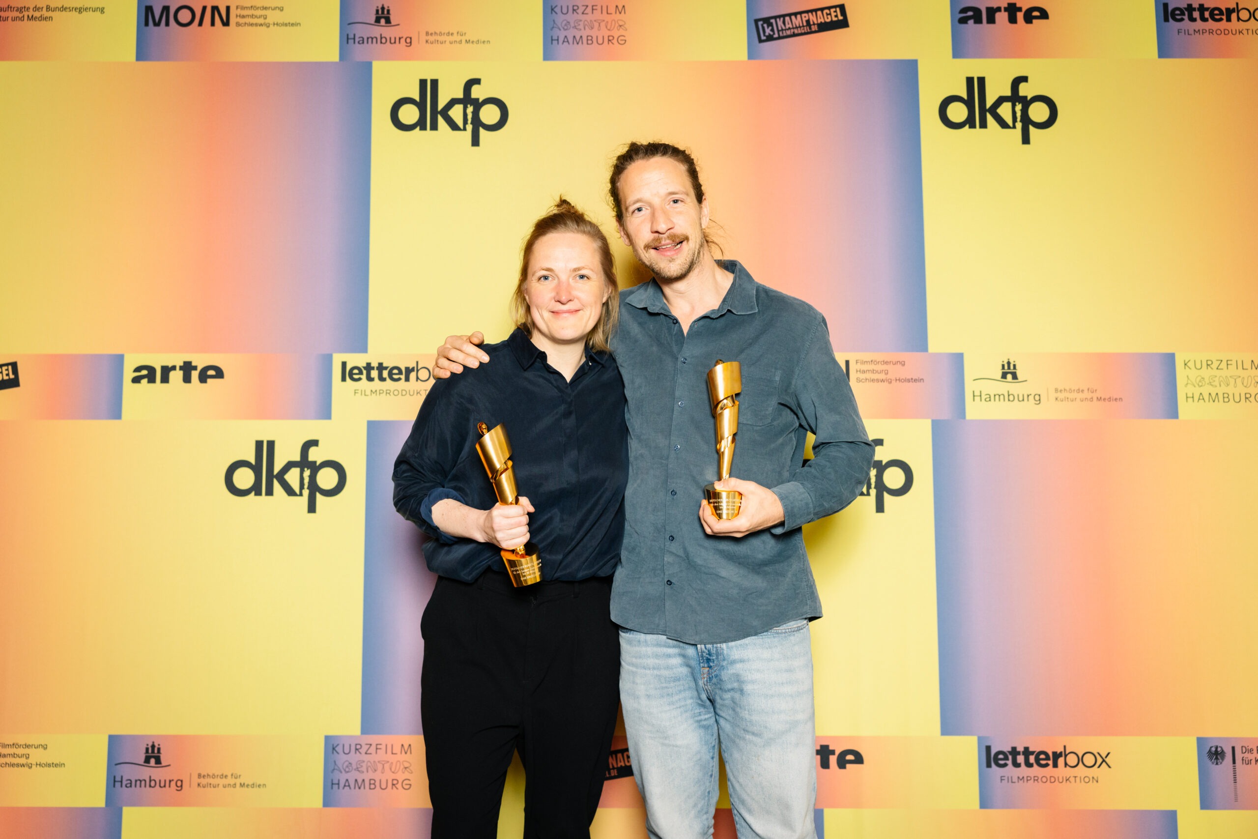 Zwei weiße Personen vor einer orang-gelben Wand, Frau und Mann. Aufschrift: dkfp. Sie halten beide einen Pokal in der Hand und lächeln.