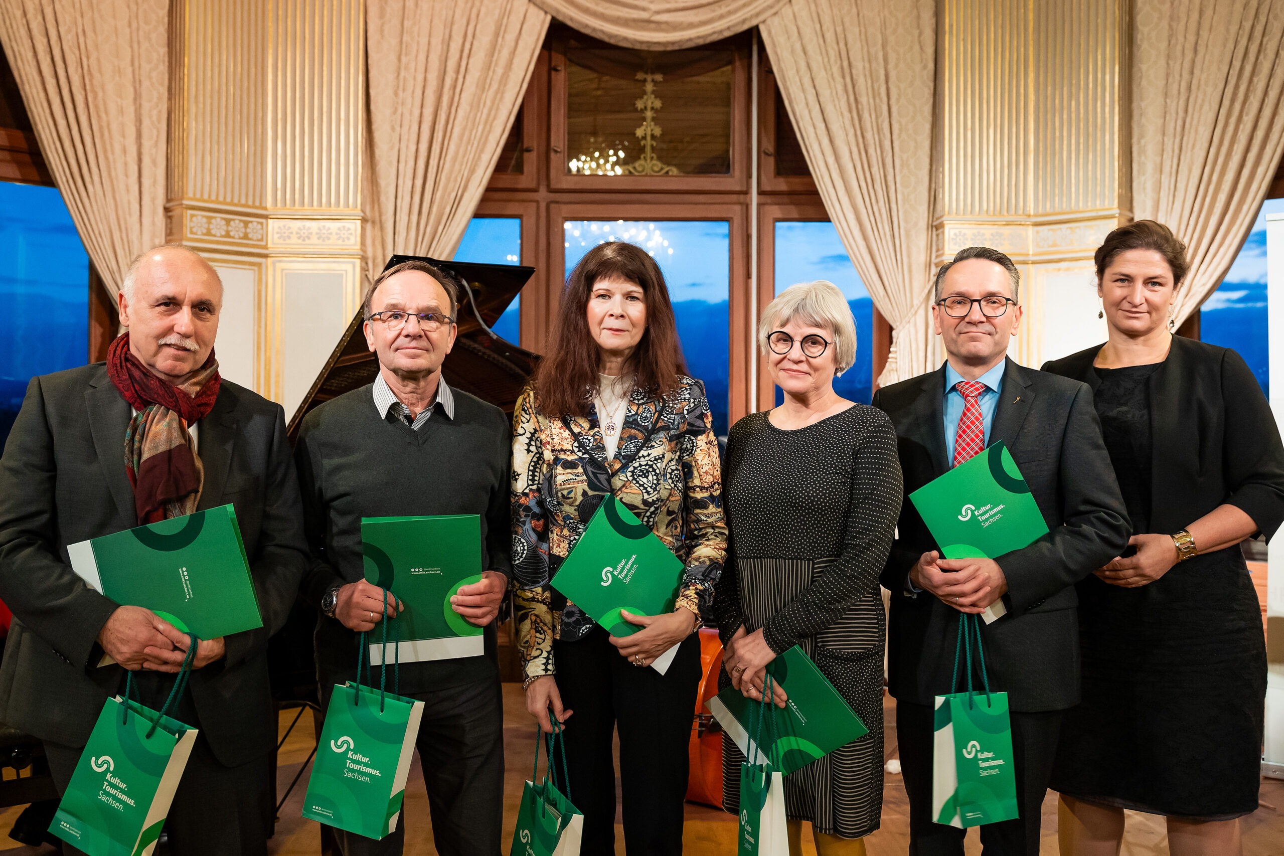 Sechs weiße Personen, drei Frauen, drei Männer, alle schick gekleidet. Fünf Menschen halten eine grüne Urkunde und grüne Taschen in der Hand mit der Aufschrift: Kultur Tourismus Sachsen.