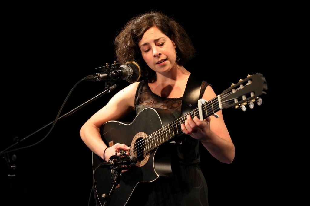 Eine weiße Frau in schwarzer Kleidung vor schwarzem Hintergrund, sie trägt eine Gitarre in den Händen und singt.
