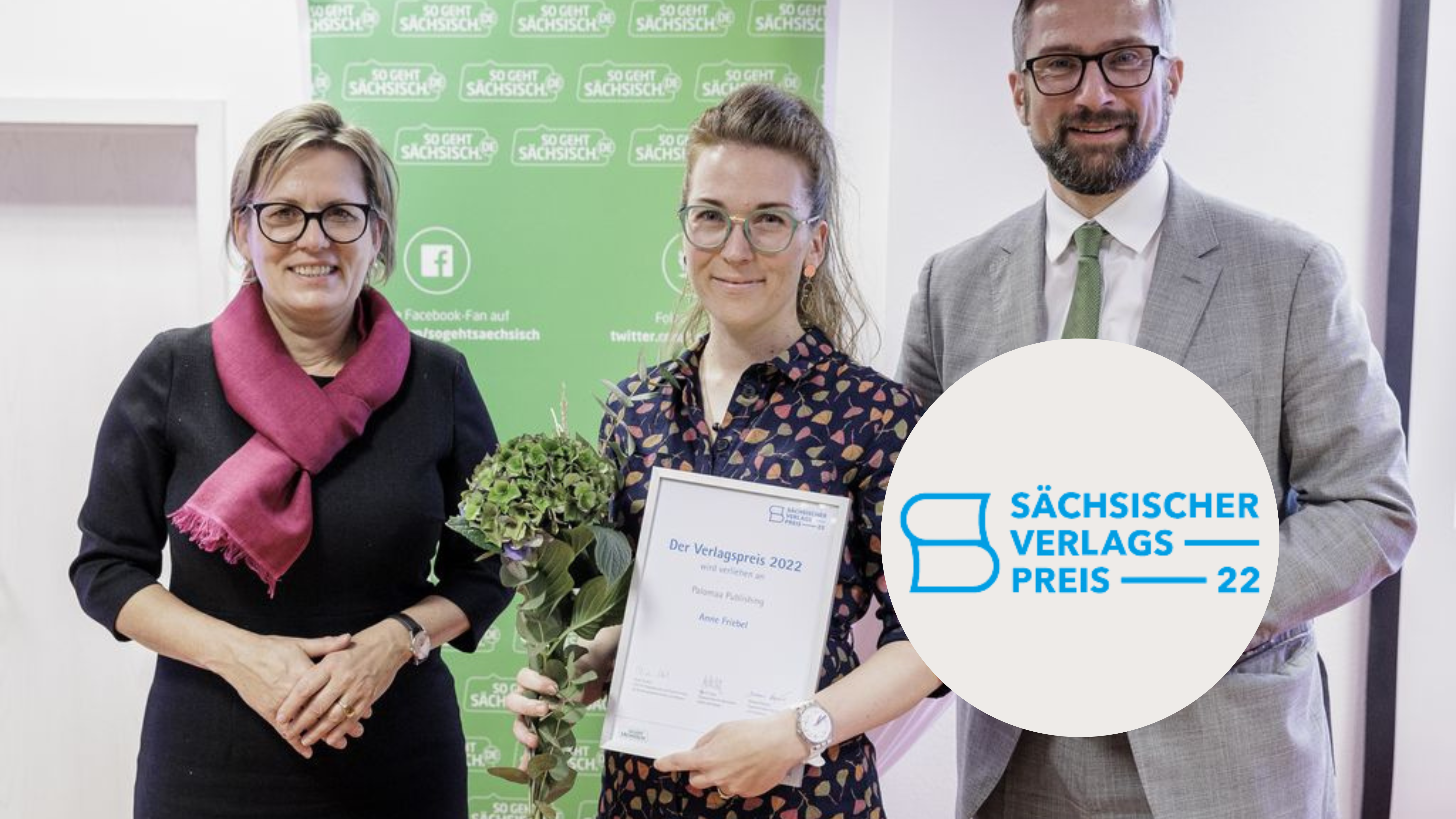Drei weiße Personen, zwei Frauen links, ein Mann rechts. Die Frau in der Mitte trägt Blumen und enie Urkunde in der Hand. Aufschrift: Sächsischer Verlagspreis 22