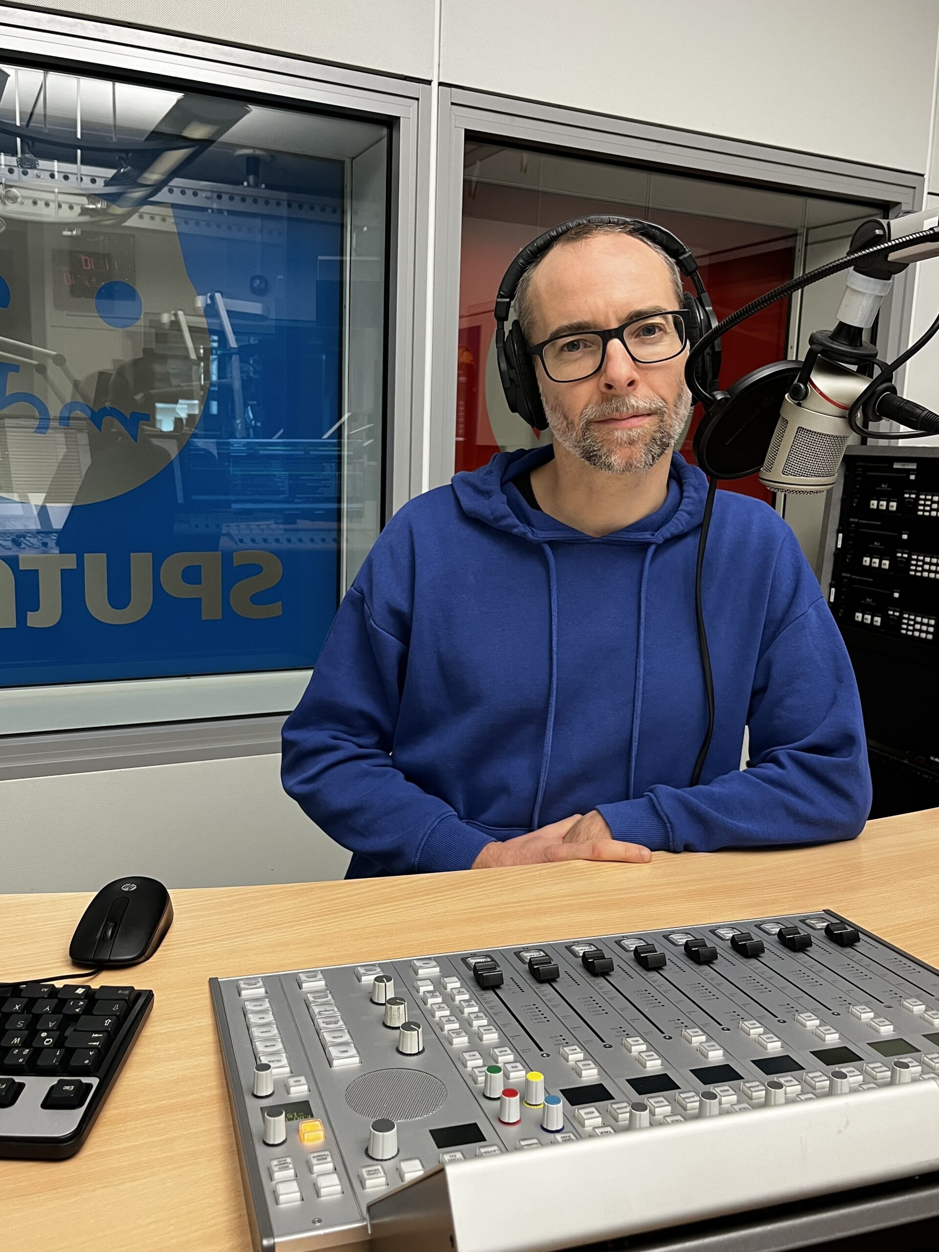 Ein Mann mit Bart und Brille in einem blauen Hoodie sitzt in einem Radio Aufnahmestudio mit Kopfhörern und Mikro.
