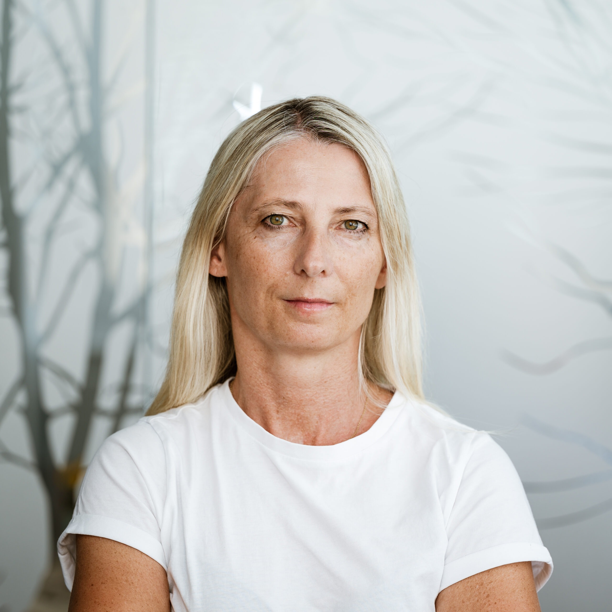 Blonde Frau mittleren Alters in einem weißen T-Shirt, im Hintergrund Nebel und kahle Bäume.