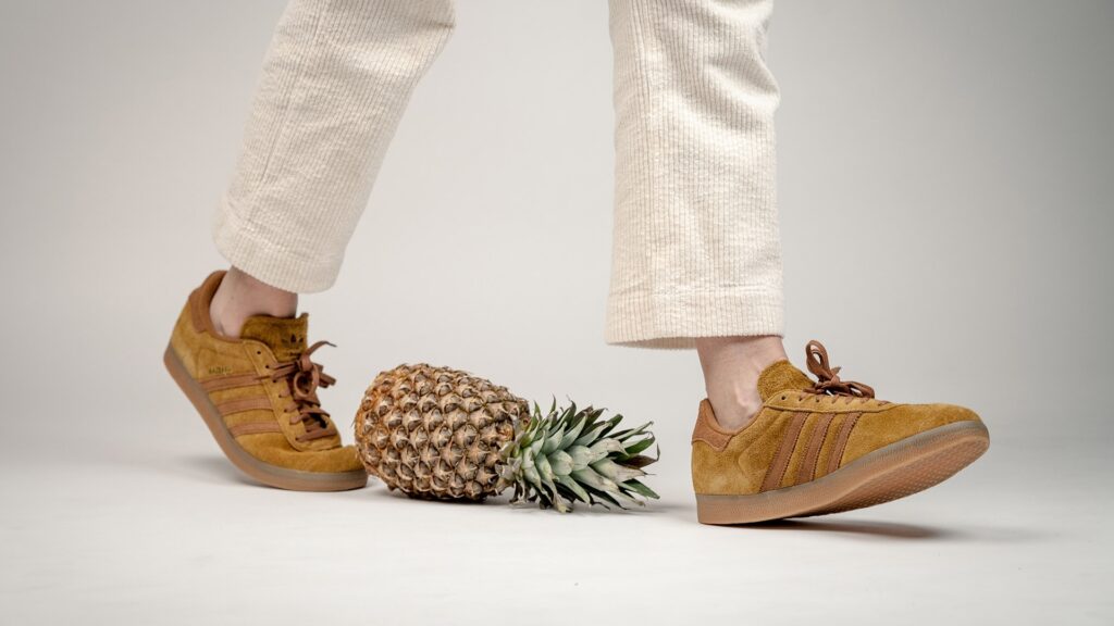 Eine Person mit weißer Hose und braunen Schuhen läuft um eine Ananas herum