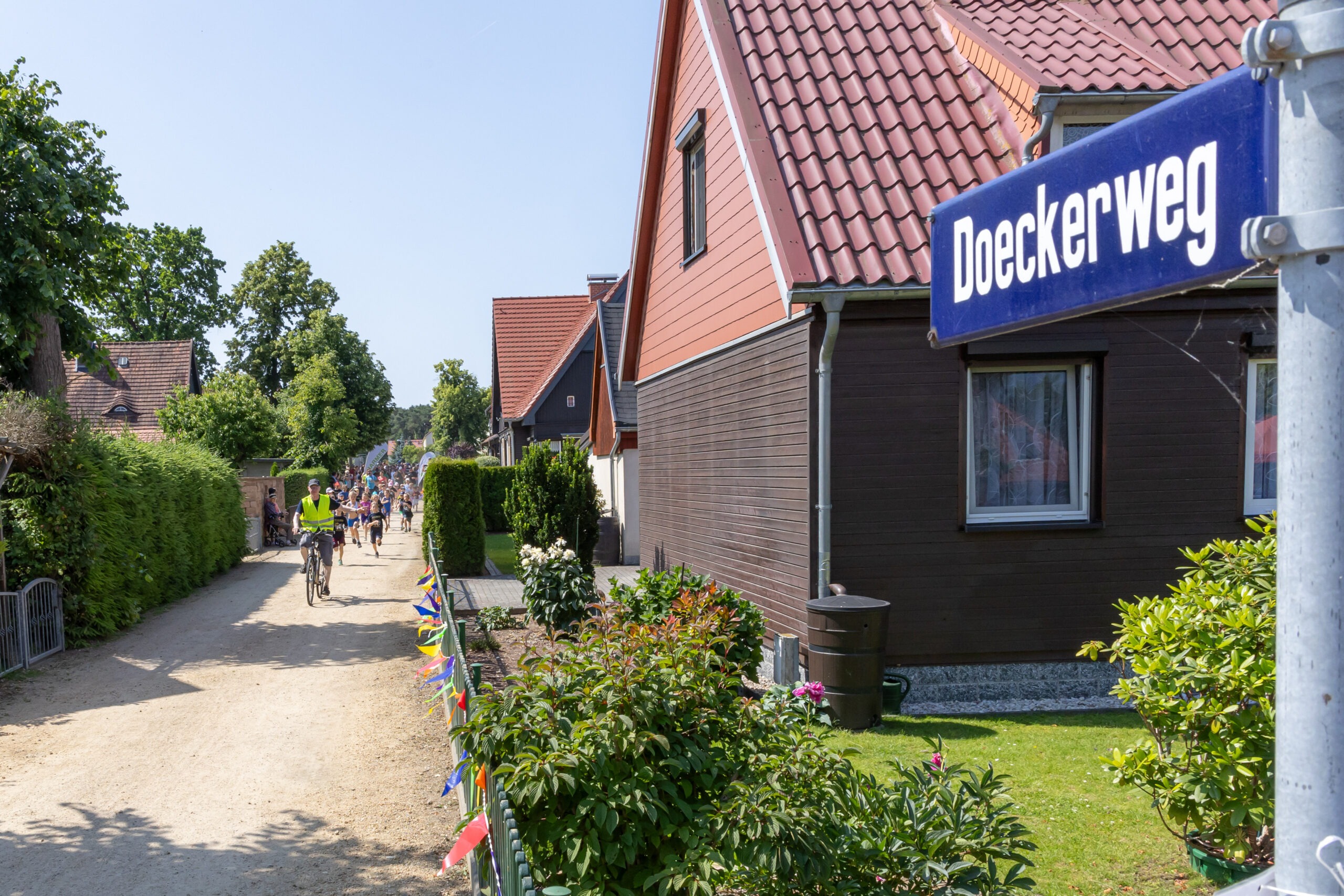 Ein Weg in einem Dorf, rechts ein Straßenschild mit der Aufschrift Doeckerweg, links eine Gruppe Fahrradfahrer