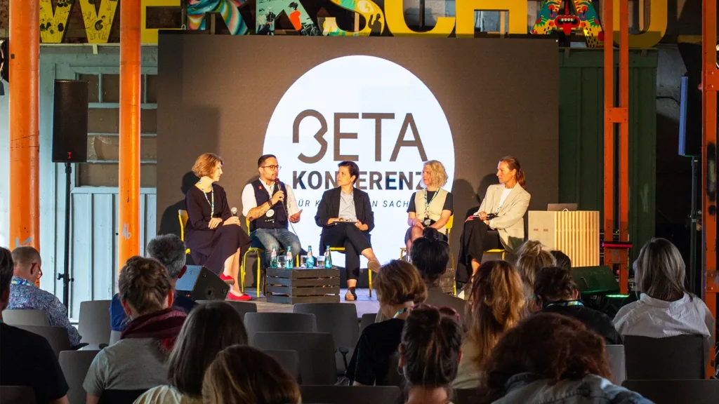 BETA Konferenz, Menschen sitzen im Podium auf einer Bühne im Hintergrund das BETA Konferenz Logo