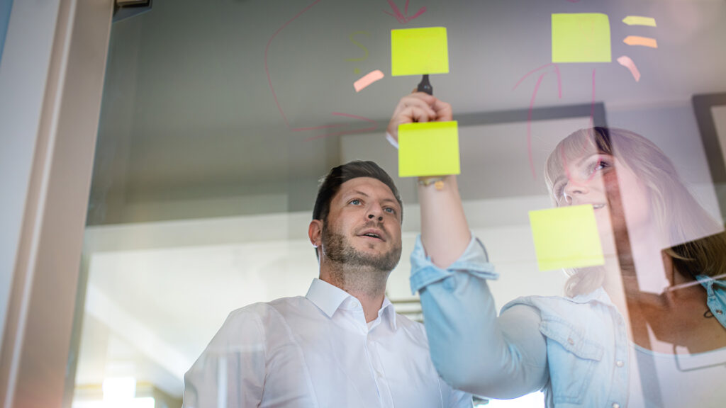 Zwei Menschen kleben glebe Klebezettel an eine Glaswand.