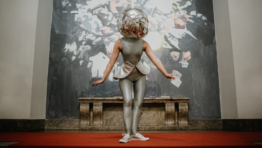 silber gekleidete Frau mit Diskokugel als Kopf auf einem roten Teppich